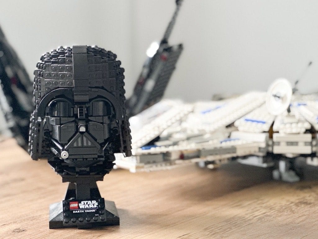 A LEGO model of Darth Vader's head with a plaque that reads "LEGO Star Wars Darth Vader," in focus in the foreground, with a blurry LEGO Star Wars spaceship model in the background on a wooden surface.